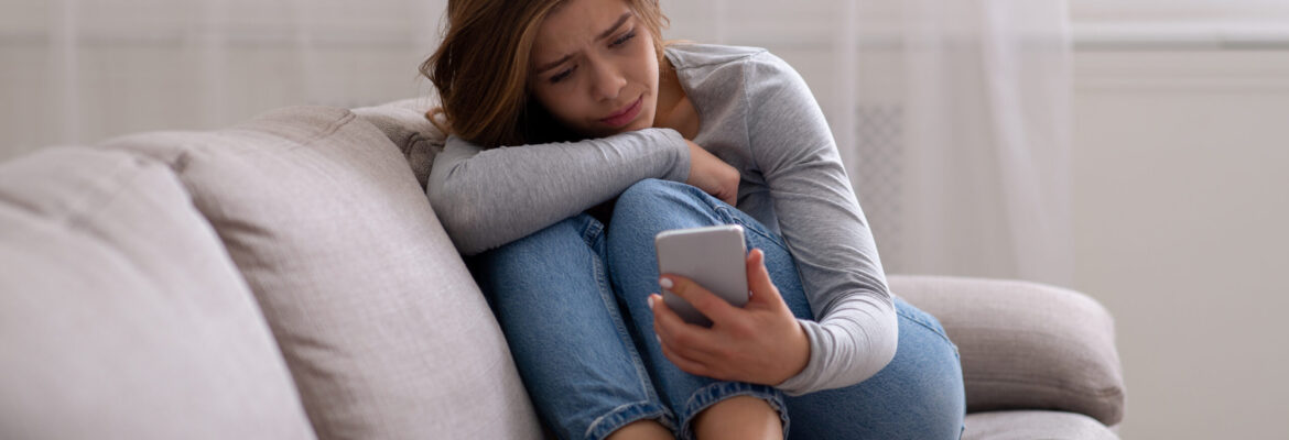 Portrait of sad young woman looking at her smartphone, suffering from cyber bullying or unhappy relationship, going through bad breakup. Depression and emotional problems concept